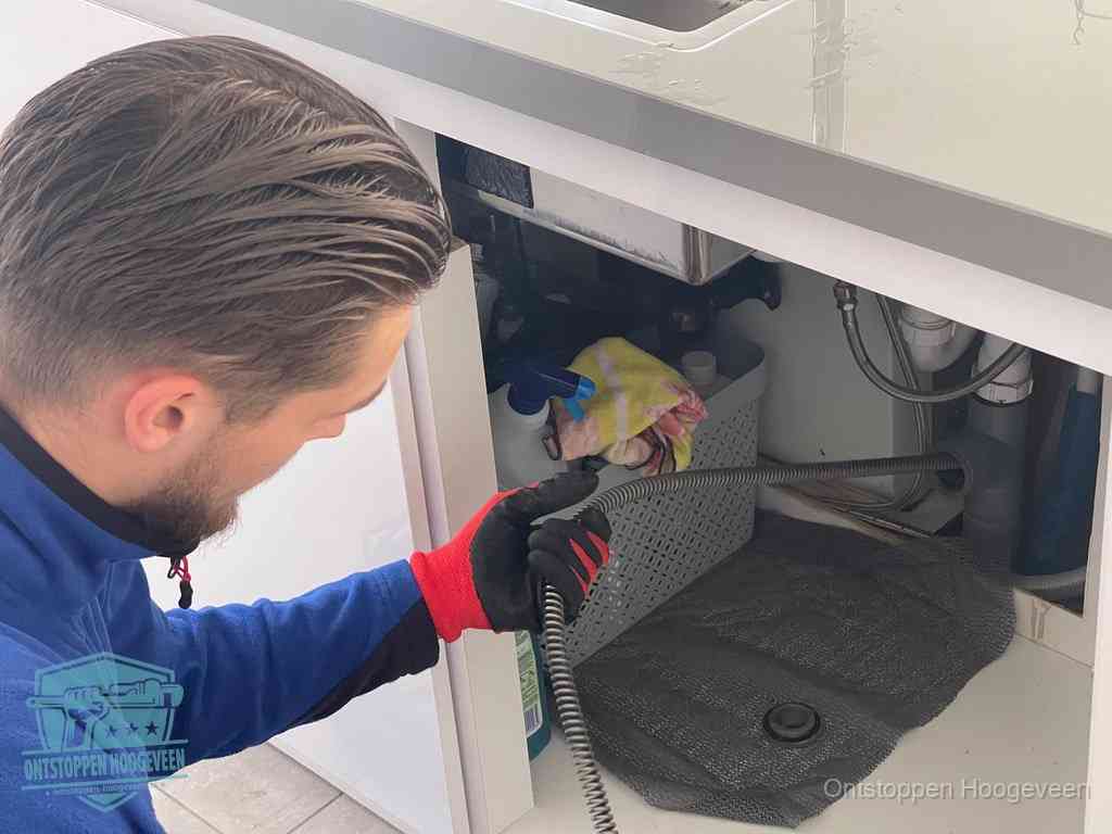 Een monteur voert een flexibele ontstoppingsveer in onder het keukenkastje, langs de sifon, om de gootsteen te ontstoppen in Hoogeveen