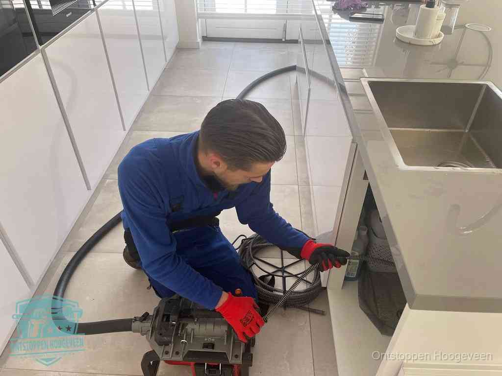 Een monteur ontstopt de keukenafvoer onder de spoelbak met een elektrische veermachine in Hoogeveen