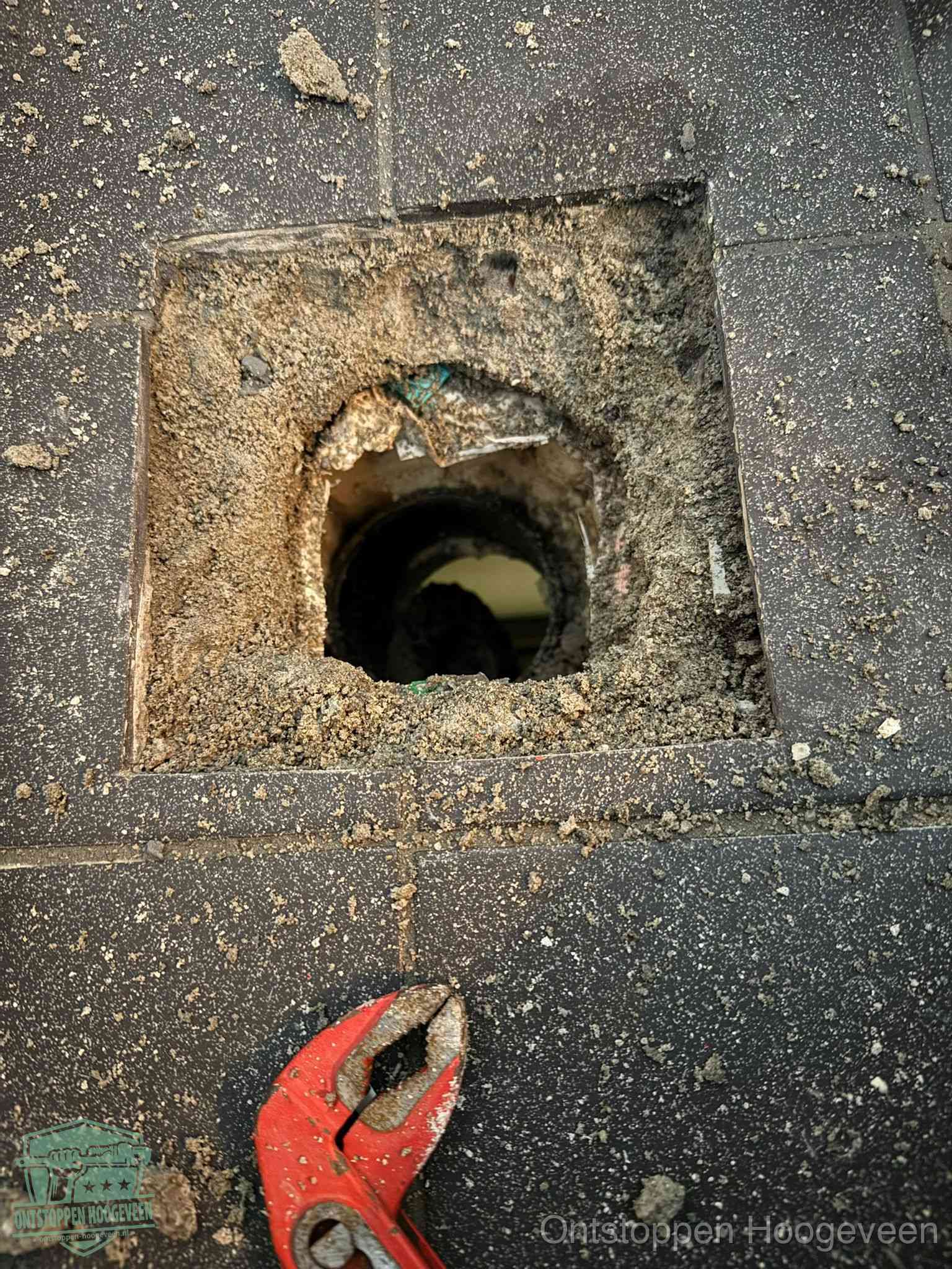 Een geopend rioolputje in een betontegelvloer met losgemaakt rooster en een zichtbare ronde afvoerbuis, het achterhalen waar de afvoerverstopping zit in Hoogeveen