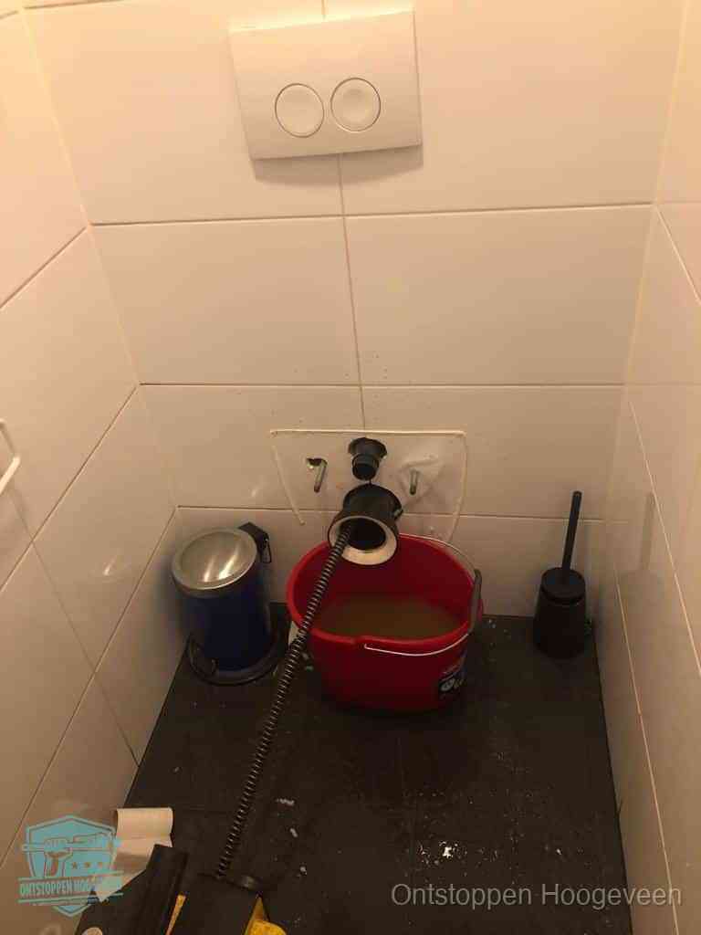 Een ontstopte wand-wc met verwijderde toiletpot waar een ontstoppingsveer in de wanduitgang verdwijnt, het achterhalen waar de verstopping zit in Hoogeveen