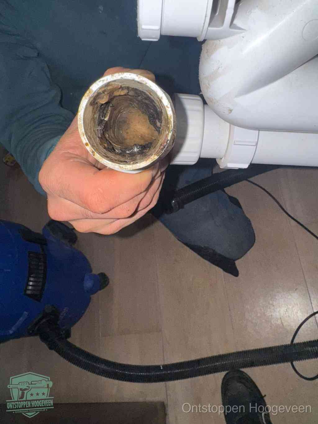 Een hand houdt een losgekoppeld PVC-afvoerbuisstuk vast dat van binnen volledig is dichtgeslibd met vet, vuil en aanslag, de meest voorkomende oorzaak van een verstopte afvoer in Hoogeveen