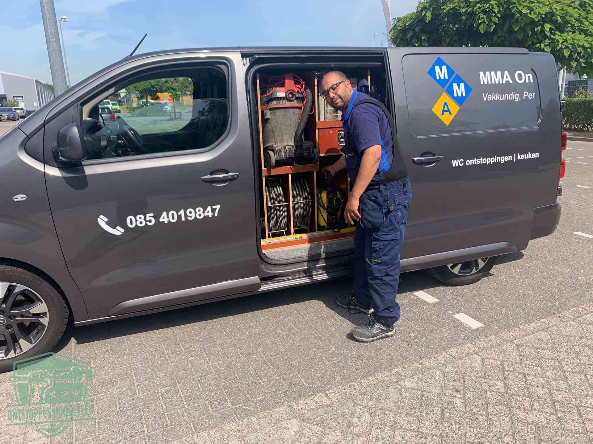 Een monteur bij de open schuifdeur van zijn bedrijfsbus op een parkeerplaats, klaar om de oorzaak van terugkerende afvoerverstoppingen aan te pakken in Hoogeveen