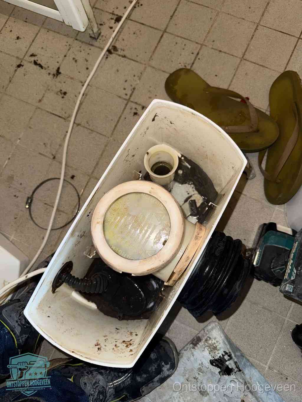Een gedemonteerde toiletpot op de vloer met ontstoppingsveer en gereedschap ernaast, het verwijderen van een vastzittend voorwerp uit de wc in Hoogeveen