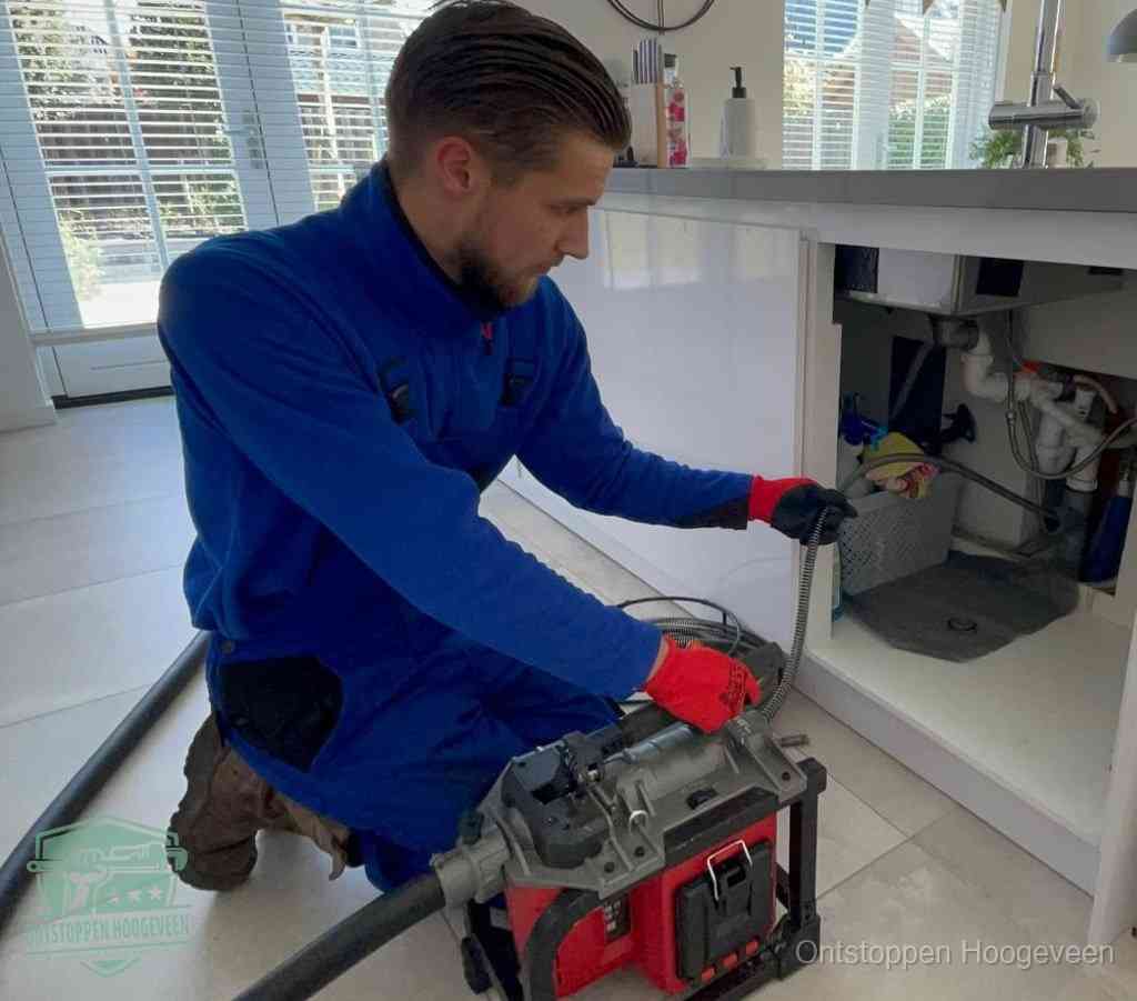 Een monteur met rode handschoenen knielt in een keuken en voert een elektrische ontstoppingsveer in tijdens het professioneel afvoer ontstoppen in Hoogeveen