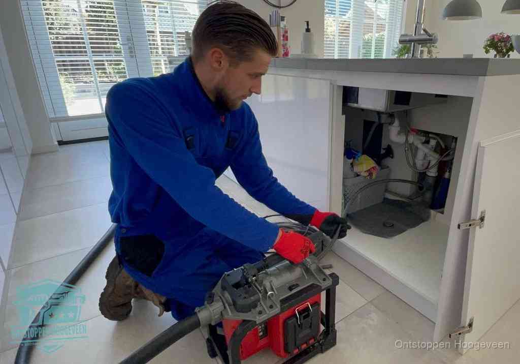 Een monteur voert een professionele elektrische ontstoppingsmachine in de afvoer onder de spoelbak tijdens het gootsteen ontstoppen in Hoogeveen